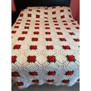May include: A white crocheted throw blanket with red and green roses. The blanket is made with a delicate stitch pattern and has a fringed edge.