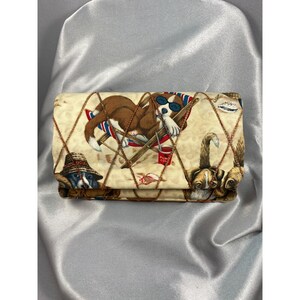 Puede incluir: Una cartera de tela beige con un estampado repetido de perros relajándose en una playa. Los perros se representan en varias poses, algunos con gafas de sol y sombreros. La cartera está doblada y asegurada con un patrón cruzado marrón.