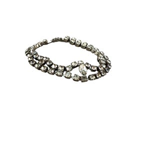 Puede incluir: Una pulsera plateada con dos filas de brillantes cuadrados transparentes. Un brillante ovalado más grande está centrado en la pulsera. La pulsera está sobre un fondo blanco.