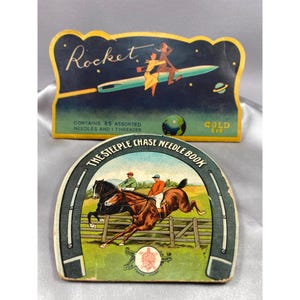 Könnte beinhalten: Zwei antike Nadelbücher. Das erste Buch ist blau mit Sternen und einem Raketenschiff, auf dem Menschen reiten. Der Text lautet "Rocket" und "Enthält 85 verschiedene Nadeln und 1 Nadelfäden. Goldenes Auge". Das zweite Buch ist grün und zeigt ein Hufeisen mit einer Pferderenn-Szene darin. Der Text lautet "The Steeple Chase Needle Book".
