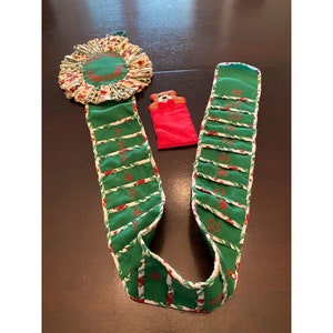 May include: A green and white fabric advent calendar with a red felt pocket and a small embroidered bear. The calendar has numbers 1 through 24 printed on it. The top of the calendar is a white and green fabric circle with the word "Christmas" embroidered on it.