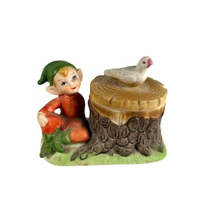 Puede incluir: Figura de cerámica que representa un elfo sentado con un atuendo rojo y detalles verdes, junto a un tocón de árbol con una paloma blanca. La figura tiene una cara pintada y está sobre una base verde.