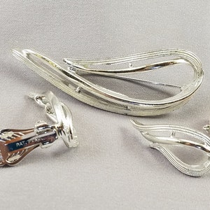 Puede incluir: Un broche y unos pendientes de clip de tono plateado. El broche y los pendientes tienen forma de lágrima con un diseño de doble línea. La parte posterior de los pendientes está marcada con "PAT. PEND."