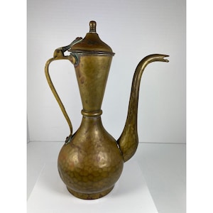Puede incluir: Una jarra de latón vintage con un pico largo y curvo y un asa. La jarra tiene una base bulbosa y una textura martillada. La tapa está sujeta con una bisagra. La jarra es de color dorado antiguo cálido.