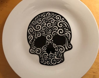 Puede incluir: Plato de cerámica blanca con un diseño de calavera negra. La calavera está decorada con intrincados patrones de remolinos. El plato es redondo y tiene un acabado liso y brillante. Un artículo decorativo para Halloween o el Día de los Muertos.