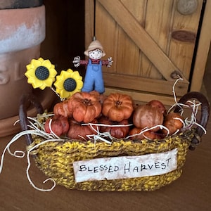 May include: A decorative basket filled with miniature orange pumpkins, straw, and a small scarecrow figurine. The basket is yellow and brown with the words "Blessed Harvest" on a white label. Two yellow sunflowers are attached.