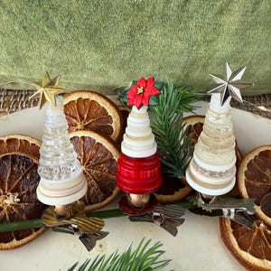 Puede incluir: Tres adornos navideños en miniatura con forma de árbol. Los árboles están hechos de botones apilados y cuentas de vidrio, rematados con adornos de estrella. Un árbol es rojo y los otros son transparentes. Los adornos están sujetos a una base con rodajas de naranja secas.