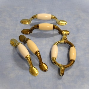 May include: Five brass cabinet knobs with white ceramic inserts. The knobs are in various shapes, including round, oval, and rectangular.