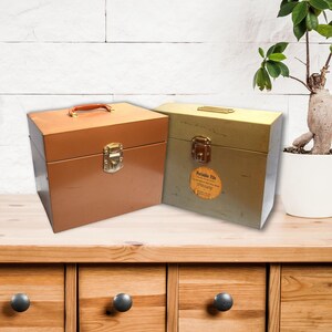 May include: Two vintage metal file boxes, one brown and one green, with a brass latch and handle. The boxes are sitting on a wooden table with drawers.