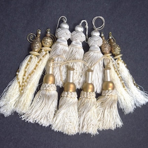 May include: Set of seven white tassel ornaments with gold accents. The tassels are made of a soft, fluffy material and have a beaded fringe. The gold accents are in the shape of small, round balls.