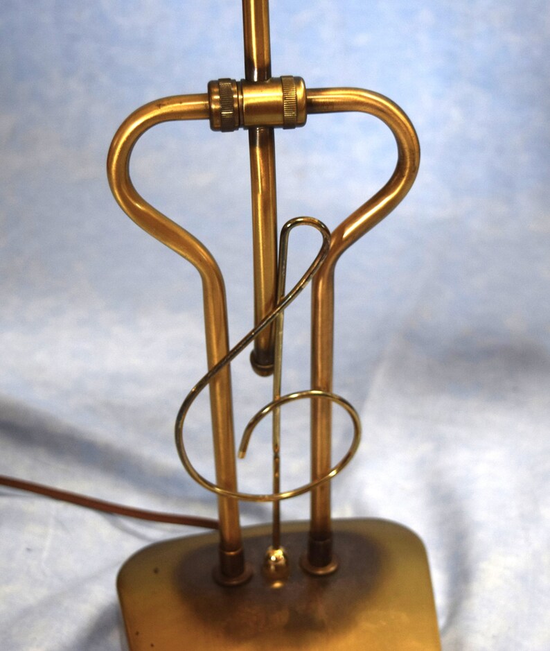 K&ouml;nnte beinhalten: Eine goldfarbene Tischlampe mit einem einzigartigen Design in Form eines Violinschl&uuml;ssels. Die Lampe hat einen runden Sockel und einen schlanken, gebogenen Schaft.