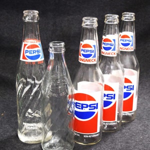 May include: Five glass Pepsi bottles, four with long necks and one with a shorter neck. The bottles are clear and have a textured surface. The bottles have red, white, and blue labels with the word "Pepsi" and "Longneck" printed on them.