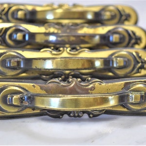 May include: A set of eight antique brass cabinet door pulls with ornate detailing. The pulls are in the shape of a long oval with a raised center section and decorative scrollwork on either end.