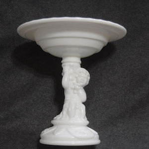 May include: White milk glass pedestal bowl with a cherub figure holding the bowl. The bowl is round and has a scalloped edge. The cherub is sculpted in detail and has a cherubic face.