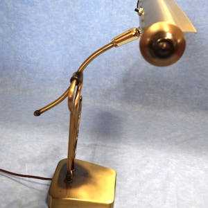 K&ouml;nnte beinhalten: Eine goldfarbene Schreibtischlampe mit einem langen, gebogenen Arm und einem rechteckigen Sockel. Die Lampe hat einen zylindrischen Schirm mit wei&szlig;em Innenraum.