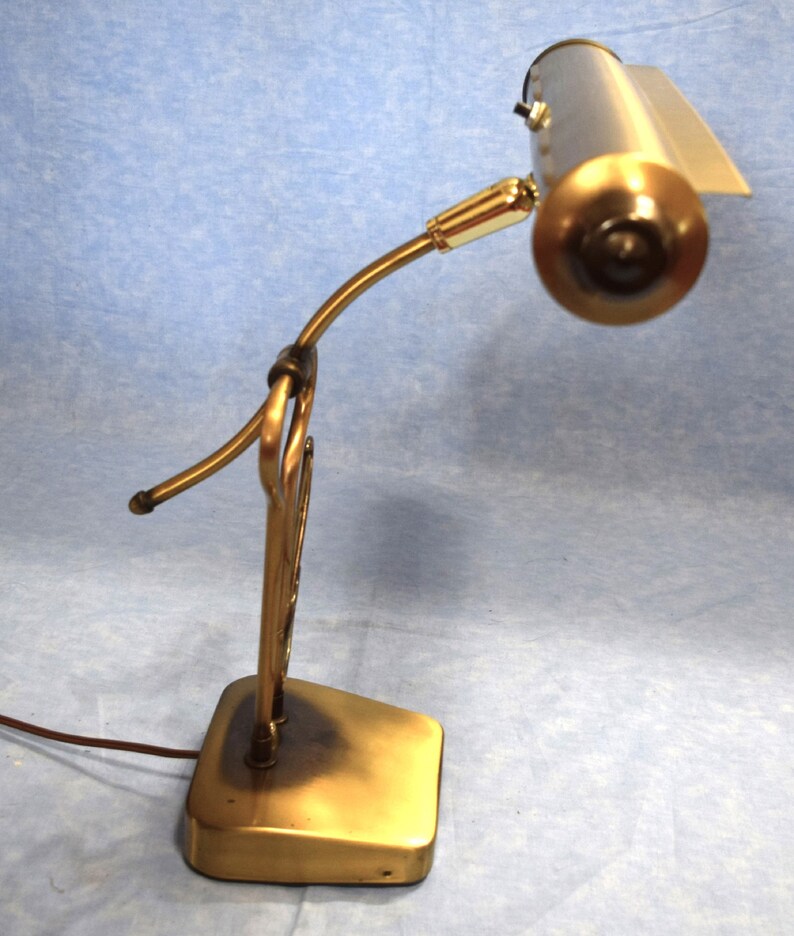 K&ouml;nnte beinhalten: Eine goldfarbene Schreibtischlampe mit einem langen, gebogenen Arm und einem rechteckigen Sockel. Die Lampe hat einen zylindrischen Schirm mit wei&szlig;em Innenraum.