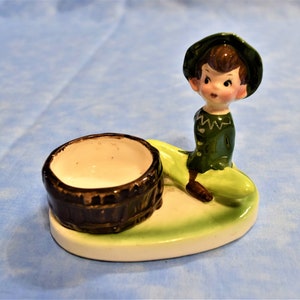 May include: A ceramic figurine of a green-clad elf sitting on a green leaf with a brown barrel. The elf is wearing a green hat and has brown hair. The figurine is on a white oval base.