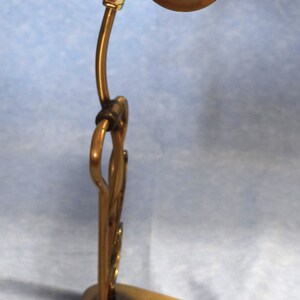 K&ouml;nnte beinhalten: Eine goldfarbene Schreibtischlampe mit einem langen, gebogenen Arm und einem zylindrischen Schirm. Der Schirm hat eine messingfarbene Oberfl&auml;che und einen schwarzen Schalter an der Seite.