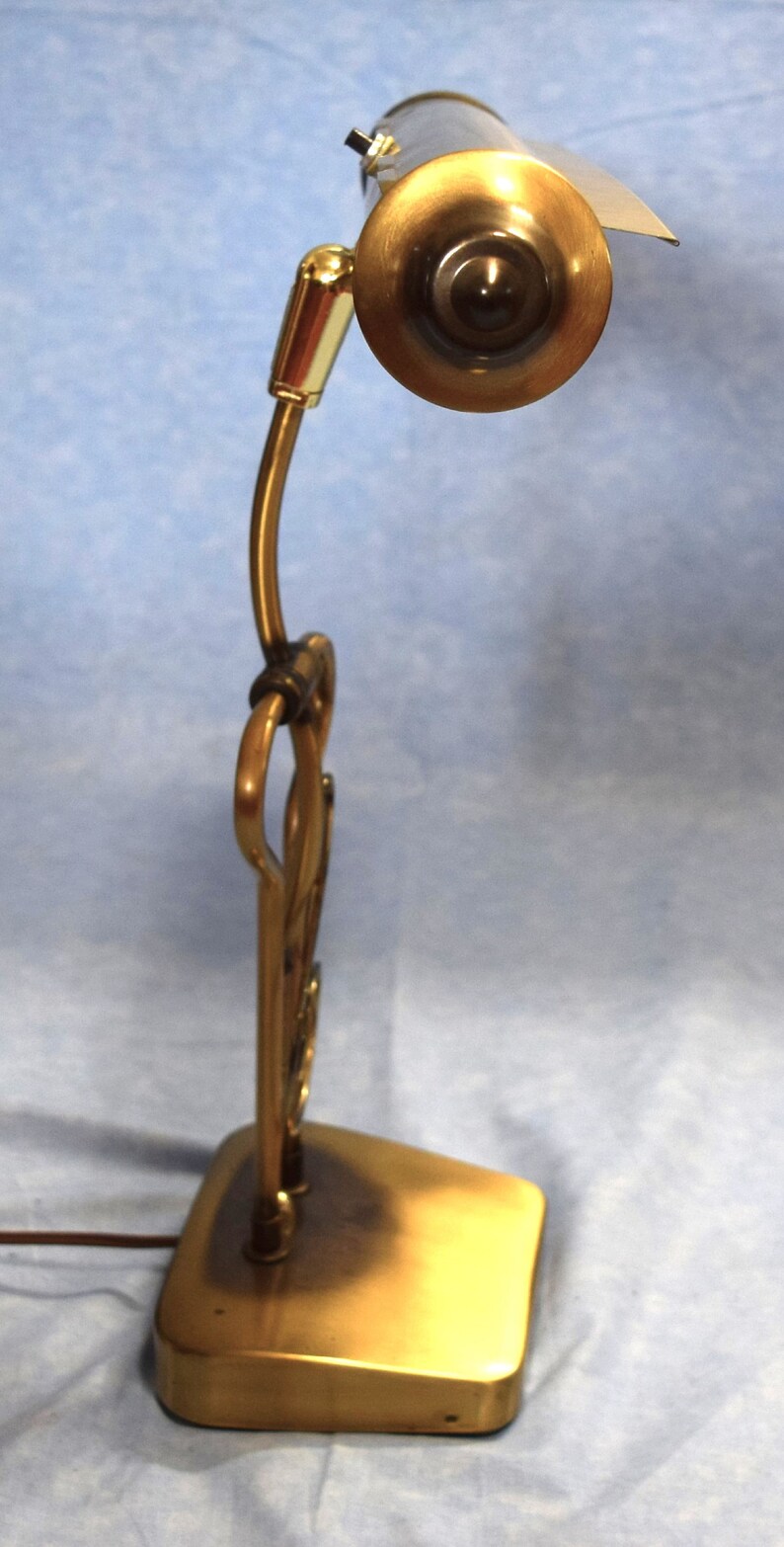 K&ouml;nnte beinhalten: Eine goldfarbene Schreibtischlampe mit einem langen, gebogenen Arm und einem zylindrischen Schirm. Der Schirm hat eine messingfarbene Oberfl&auml;che und einen schwarzen Schalter an der Seite.