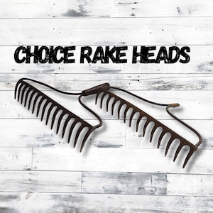 May include: Two antique metal rake heads with curved tines, displayed against a white and gray wooden background. The text "CHOICE RAKE HEADS" is printed in black above the rakes. These garden tools are likely for yard work.