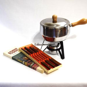 May include: A stainless steel fondue pot with a wooden handle and lid knob, resting on a black metal stand with a burner. Beside it is a rectangular box labeled 6 FONDUE FORKS containing six stainless steel forks with brown wooden handles.