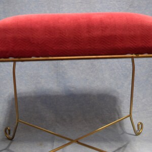 May include: A red velvet cushioned footstool with a gold metal frame. The frame has a simple design with curved legs.