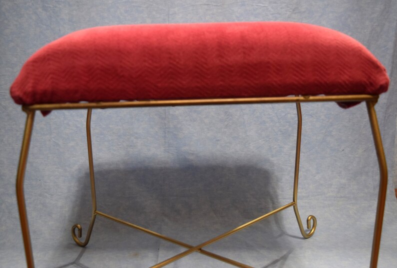 May include: A red velvet cushioned footstool with a gold metal frame. The frame has a simple design with curved legs.