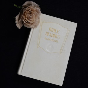 May include: A white book with the title "Bible Readings for the Home" on the cover. A beige fabric rose is on top of the book.