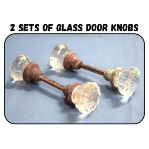 May include: Two sets of glass door knobs with clear, faceted, flower-shaped glass handles and rusted metal spindles and bases. The text 2 SETS OF GLASS DOOR KNOBS is printed above the image. The knobs feature a classic, multi-sided design.