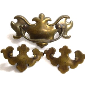 May include: Three brass drawer pulls with a metallic gold finish. One pull features a curved, swinging handle attached to a decorative backplate, while the other two are matching backplates without handles, all displaying ornate, symmetrical shapes.