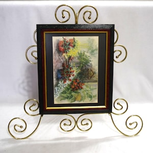 May include: A framed watercolor painting of a garden scene, displayed on a decorative gold-colored metal stand. The frame is black with a gold inner border. The stand features ornate scrollwork.