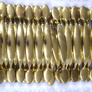 May include: A collection of numerous gold-colored metal cabinet pulls arranged in a dense, side-by-side formation. Each pull features a smooth, elongated, spindle-like shape with a reflective, polished finish and rounded ends for mounting.
