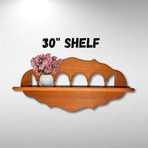 May include: A 30" wooden shelf with a decorative arched design and a scalloped edge. The shelf is made of light brown wood and has a white vase with pink flowers on top.