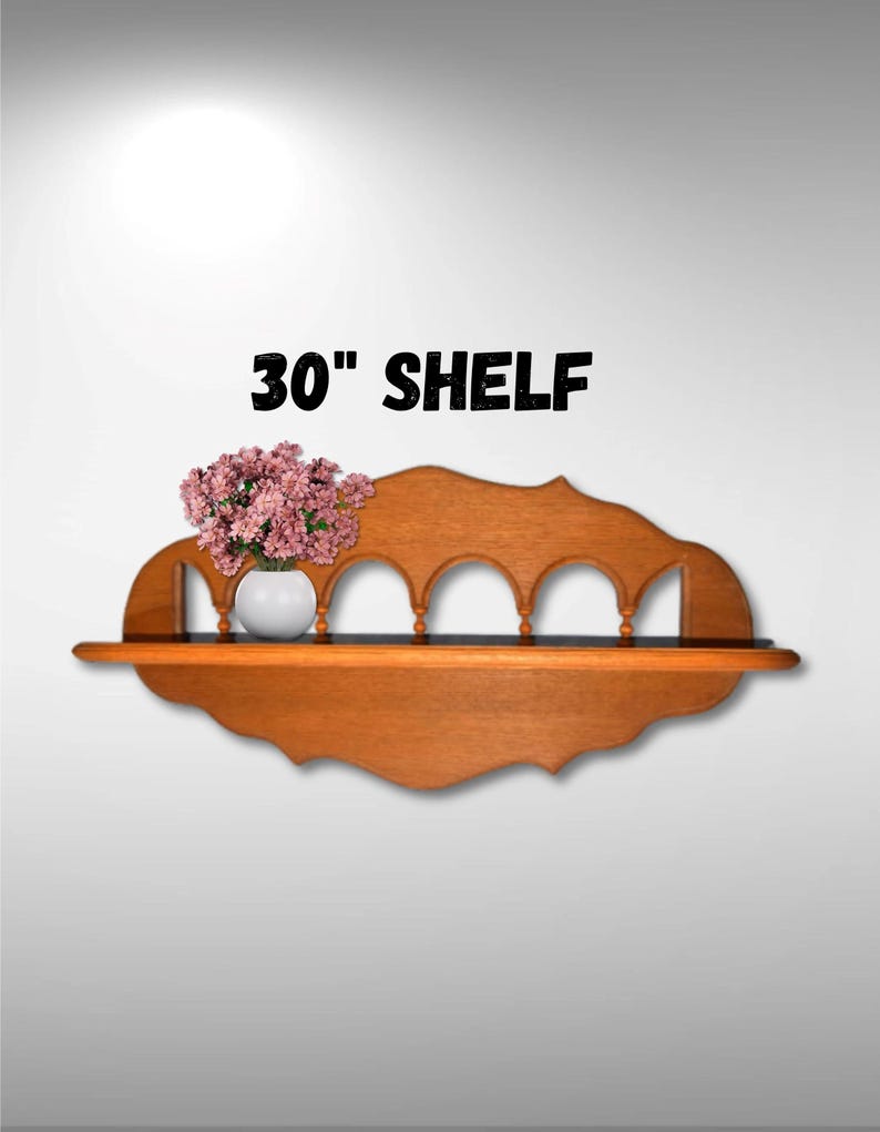 May include: A 30" wooden shelf with a decorative arched design and a scalloped edge. The shelf is made of light brown wood and has a white vase with pink flowers on top.