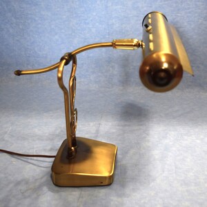 K&ouml;nnte beinhalten: Eine Schreibtischlampe aus Messing mit einem gebogenen Arm und einem langen, rechteckigen Schirm. Die Lampe hat einen dekorativen Sockel mit einem Rollendesign.