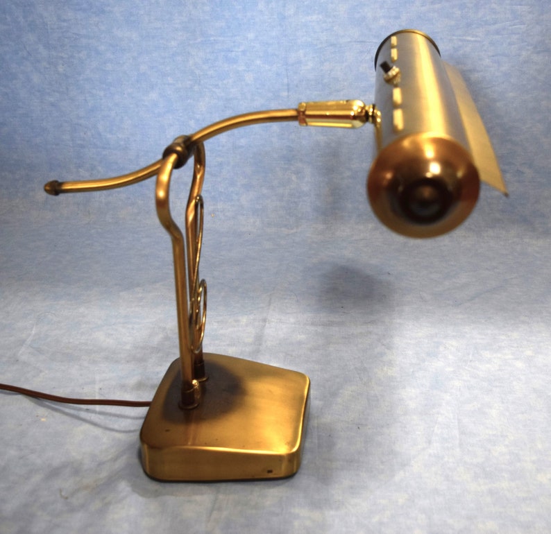 K&ouml;nnte beinhalten: Eine Schreibtischlampe aus Messing mit einem gebogenen Arm und einem langen, rechteckigen Schirm. Die Lampe hat einen dekorativen Sockel mit einem Rollendesign.
