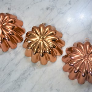 May include: Three copper-colored, flower-shaped cake molds with fluted edges. The molds have a shiny finish and are arranged in a row on a white marble surface.