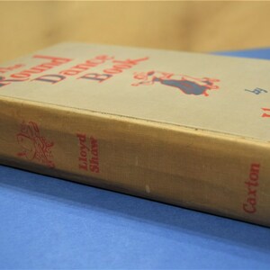 May include: The Round Dance Book by Lloyd Shaw, a vintage book with a tan cover and red text. The book features a drawing of a couple dancing.