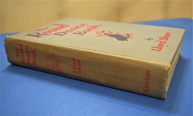 May include: The Round Dance Book by Lloyd Shaw, a vintage book with a tan cover and red text. The book features a drawing of a couple dancing.