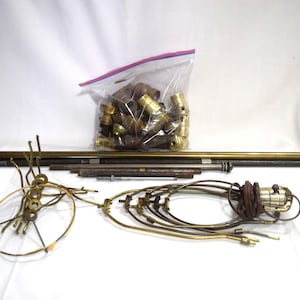 May include: A collection of brass lamp parts, including a long metal rod, a circular metal frame, and several curved metal pieces. The parts are arranged on a white surface. A plastic bag filled with additional brass parts is also visible.
