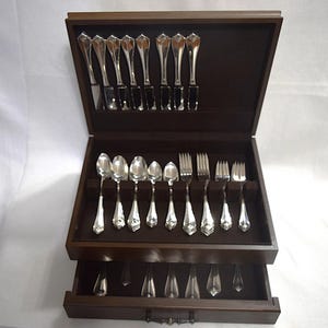 May include: A wooden box with a brown velvet interior holds a set of silverware. The silverware includes forks, knives, and spoons. The box has a drawer for additional silverware.