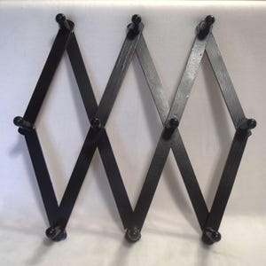 May include: Black wooden accordion-style coat rack with multiple pegs. The rack is designed with a diamond pattern and can be expanded or contracted. The pegs are small and round, providing a place to hang items. The finish is matte black.