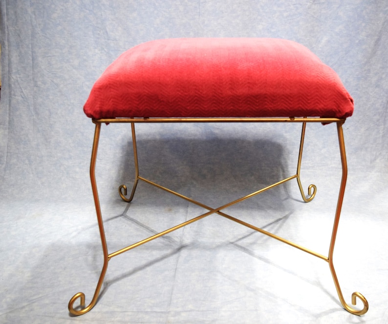 May include: A red velvet upholstered stool with a gold metal frame. The stool has four legs with a decorative scroll design at the bottom of each leg. The legs are connected by a criss-cross design.