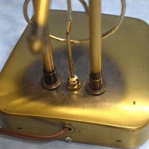 K&ouml;nnte beinhalten: Ein goldfarbener Metall-Tischlampenfu&szlig; mit einem runden Design und drei Stromkabeln. Die Basis hat eine abgenutzte, gealterte Oberfl&auml;che.