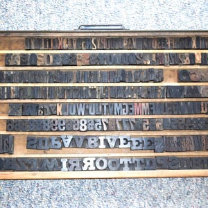 May include: A wooden printing block tray filled with rows of individual letterpress printing blocks. The blocks are made of wood and metal and feature a variety of letters, numbers, and symbols.