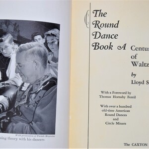 May include: A black and white photo of a man in a suit, surrounded by a group of young people. The man is writing on a piece of paper. The text on the page reads "The Round Dance Book A Century of Waltzing by Lloyd Shaw".