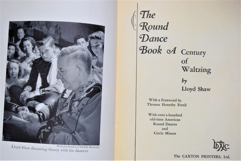 May include: A black and white photo of a man in a suit, surrounded by a group of young people. The man is writing on a piece of paper. The text on the page reads "The Round Dance Book A Century of Waltzing by Lloyd Shaw".