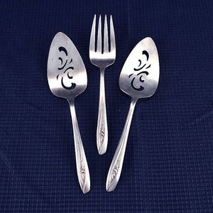 May include: Three silver cake servers with decorative cutouts. The servers have ornate handles and are on a dark blue background.