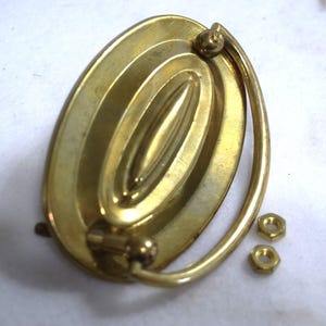 May include: A brass oval drawer pull with a curved handle and two matching nuts. The pull has a series of concentric oval ridges. The hardware is a warm gold color.