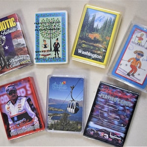 May include: A collection of eight different playing card decks. The decks feature a variety of designs, including a patriotic theme, a skyline, a race car driver, and a Washington state scene.